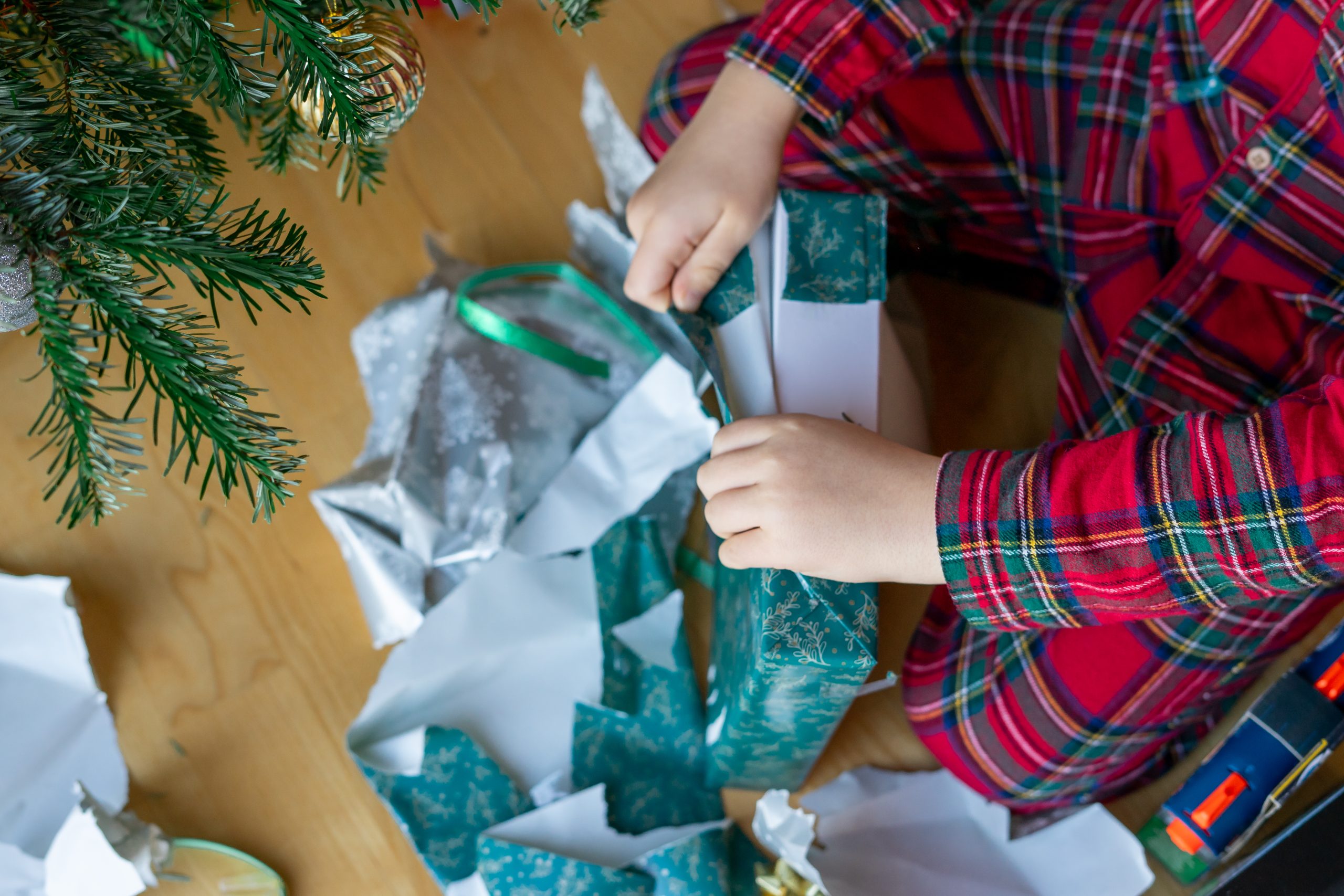 A child in red plaid pajamas opens Christmas gifts wrapped in colorful wrapping paper near the Christmas tree in the morning.