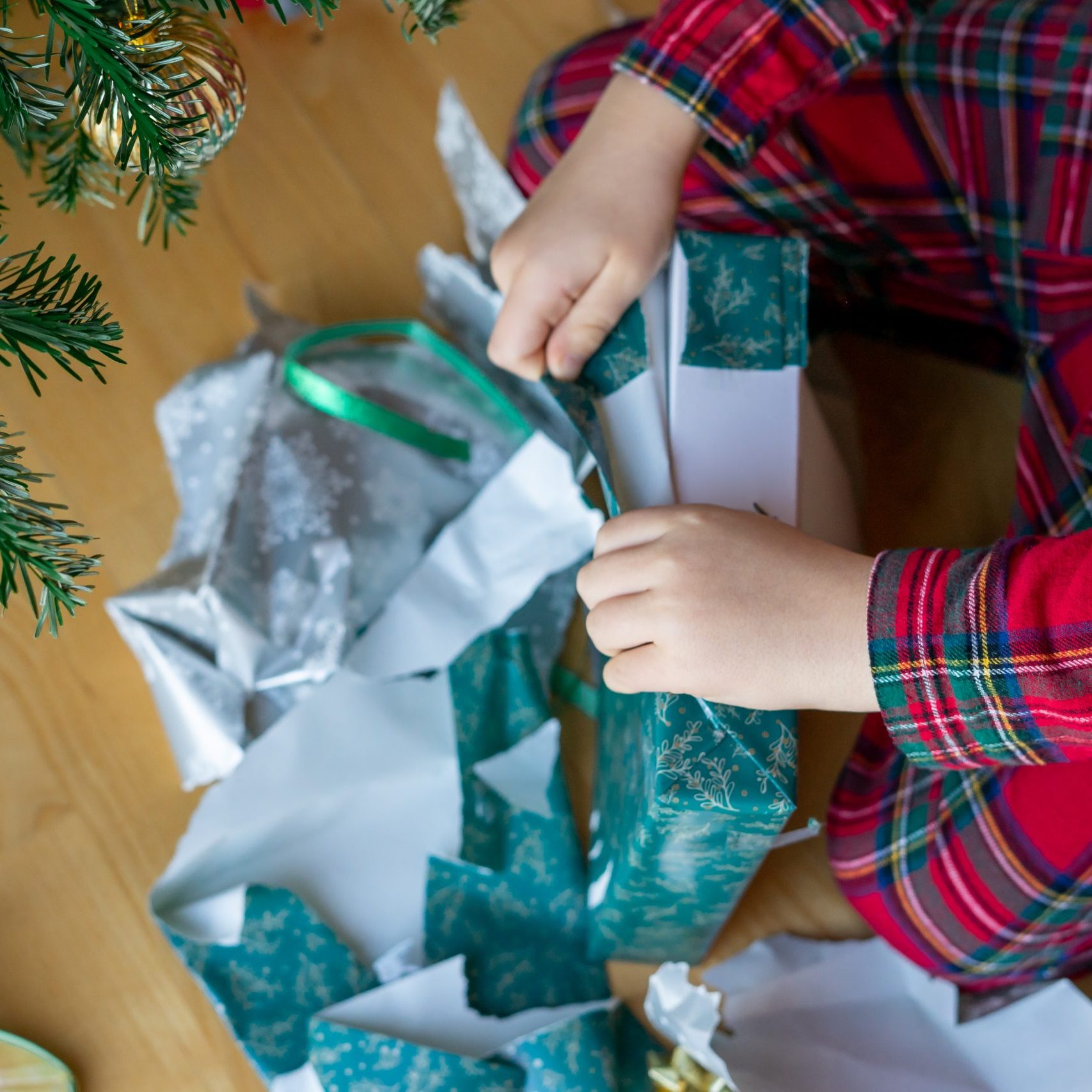 A child in red plaid pajamas opens Christmas gifts wrapped in colorful wrapping paper near the Christmas tree in the morning.